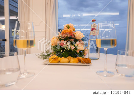 A table set with pink and peach roses, bread, white wine, and glassware. Large windows reveal a cloudy sky and the Tyrrhenian Sea in Amalfi, Italy. 131934842