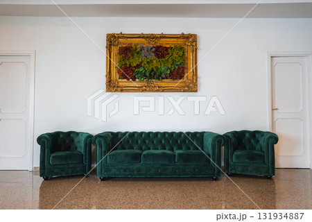 A refined room with green velvet tufted sofa and armchairs, polished stone floor, white walls, and a vibrant painting in a golden ornate frame. A refined room with green velvet tufted sofa and armchairs, polished stone floor, white walls, and a vibrant painting in a golden ornate frame. 131934887
