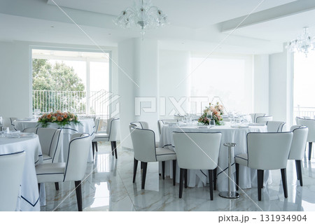Modern dining space with white walls, marble floors, round tables, floral centerpieces, and large windows offering partial views of greenery and sea. Modern dining space with white walls, marble floors, round tables, floral centerpieces, and large windows offering partial views of greenery and sea. 131934904