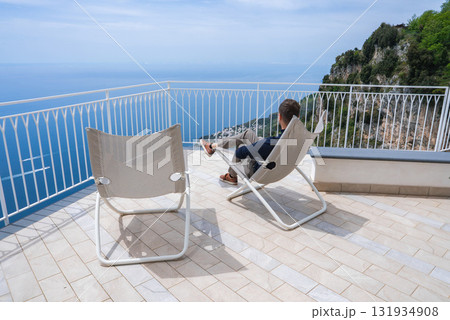 A man sits on a modern lounge chair on a tiled terrace with a white railing, overlooking the Tyrrhenian Sea and a rocky Mediterranean coastline. 131934908