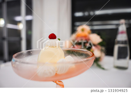 A creamy yellow dessert in a pink glass, topped with whipped cream, a red berry, and mint. Blurred floral arrangement and water bottle in background. 131934925