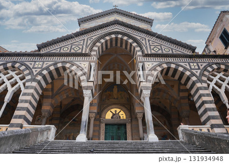 The Amalfi Cathedral features Romanesque, Byzantine, and Gothic styles, with striped arches, a golden mosaic, and a grand staircase under a clear sky. 131934948
