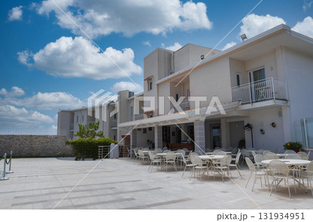A flat roofed building with balconies under a blue sky, featuring a tiled patio with white tables and chairs, greenery, and a low stone wall. 131934951