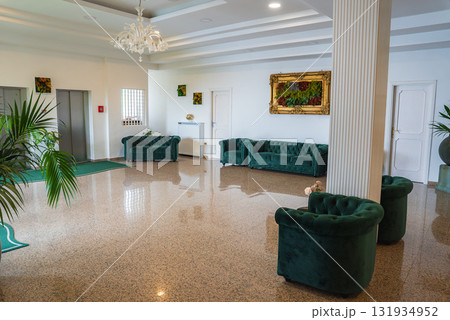 Interior of a hotel lobby in Amalfi, Italy, featuring marble flooring, green velvet seating, a chandelier, gold framed artwork, and decorative plants. 131934952