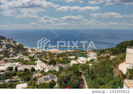 View of Capri, Italy featuring a harbor with boats, whitewashed buildings, lush greenery, a winding road, and a funicular railway under a partly cloudy sky. View of Capri, Italy featuring a harbor with boats, whitewashed buildings, lush greenery, a winding road, and a funicular railway under a partly cloudy sky. 131935012