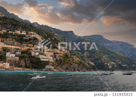 Positano's vibrant buildings cascade down a steep cliff, surrounded by greenery, mountains, and the Mediterranean Sea under a dramatic cloudy sky. 131935015