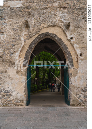 A Gothic style stone archway with green metal gates leads to a garden with trees and a pathway. People walk through, adding scale and activity. 131935025