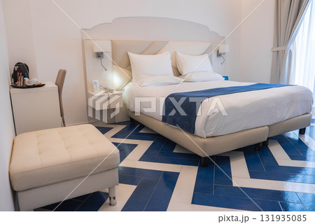 Bright hotel room featuring a double bed with white linens, blue runner, chevron patterned tiled floor, desk, coffee station, and natural light. Bright hotel room featuring a double bed with white linens, blue runner, chevron patterned tiled floor, desk, coffee station, and natural light. 131935085
