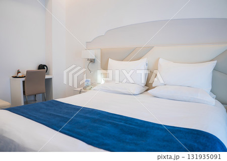 A well lit hotel room in Amalfi, Italy, featuring a neatly made bed with white linens, a blue throw, a geometric headboard, and guest amenities. 131935091
