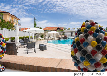 Outdoor pool with lounge chairs, umbrellas, and ceramic decorations, set against red roofed buildings, greenery, and a bright blue sky in Amalfi, Italy. 131935093