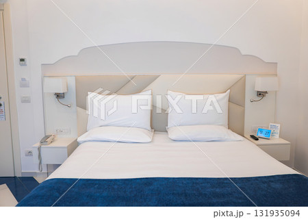 A neatly arranged hotel room featuring a double bed with white linens and a blue blanket, white walls, blue tiled flooring, and minimalistic decor. 131935094