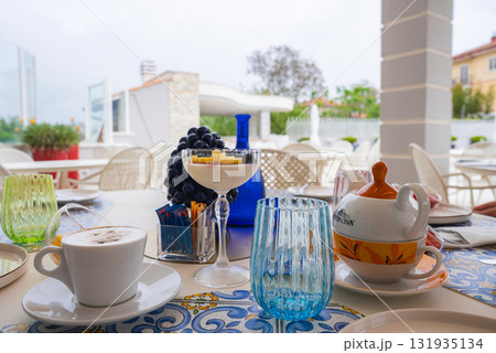 A vibrant outdoor dining table in Amalfi, Italy, featuring a cappuccino, dessert, floral teapot, colorful glassware, and lush greenery in the background. 131935134