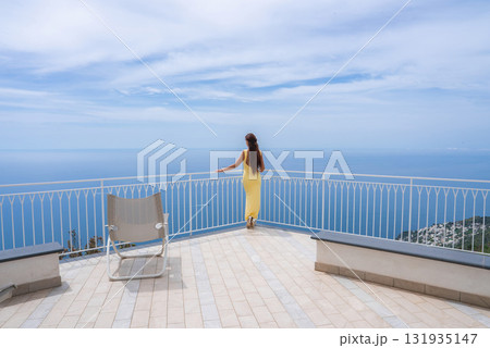 A woman in a yellow dress stands on a tiled terrace with a white railing, overlooking the Tyrrhenian Sea, coastline, and distant buildings in Amalfi. A woman in a yellow dress stands on a tiled terrace with a white railing, overlooking the Tyrrhenian Sea, coastline, and distant buildings in Amalfi. 131935147