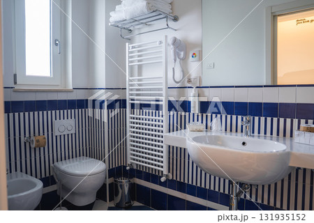 A modern bathroom featuring blue and white striped tiles, a wall mounted sink, toilet, bidet, heated towel rack, and natural light from a small window. A modern bathroom featuring blue and white striped tiles, a wall mounted sink, toilet, bidet, heated towel rack, and natural light from a small window. 131935152