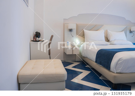 Elegant hotel room featuring a large bed with white linens, blue accents, geometric patterned tiled floor, and minimalist furnishings in Amalfi, Italy. Elegant hotel room featuring a large bed with white linens, blue accents, geometric patterned tiled floor, and minimalist furnishings in Amalfi, Italy. 131935179