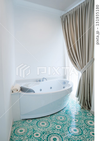 White jacuzzi style bathtub with built in jets, turquoise and white patterned tiles, and natural light through a tied back beige curtain in Amalfi. 131935180