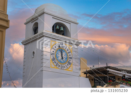 The Torre dell'Orologio in Capri, Italy, features a blue and gold clock face, domed top, and bells, set against a pink and blue sky with nearby terrace. 131935230