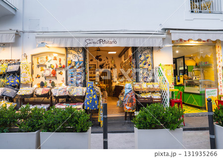 A storefront in Positano, Italy, with colorful textiles and ceramics, next to a pizzeria with a yellow and green interior, framed by white walls and greenery. 131935266