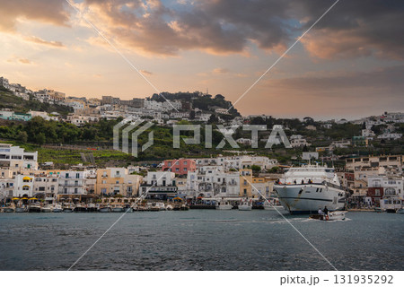 Marina Grande on Capri, Italy, features colorful waterfront buildings, a large yacht, smaller boats, and lush hills under a warm sunset sky. 131935292