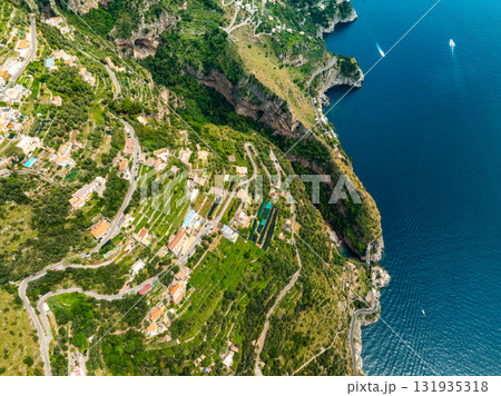 Aerial image of the Amalfi Coast in Italy, showing terraced hills, colorful buildings, winding roads, cliffs, a luxury hotel, and boats on the sea. Aerial image of the Amalfi Coast in Italy, showing terraced hills, colorful buildings, winding roads, cliffs, a luxury hotel, and boats on the sea. 131935318