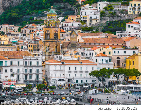 Amalfi Cathedral with its ornate bell tower and green dome stands out among pastel buildings, lush cliffs, and a vibrant waterfront promenade. Amalfi Cathedral with its ornate bell tower and green dome stands out among pastel buildings, lush cliffs, and a vibrant waterfront promenade. 131935325