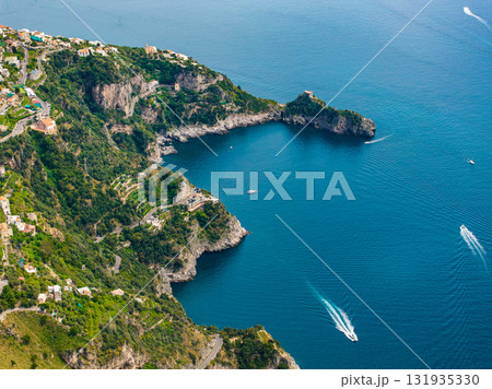 Aerial view of the Amalfi Coast in Italy, showing cliffs, green hills, a small bay, boats on the Tyrrhenian Sea, and villas along winding roads. 131935330