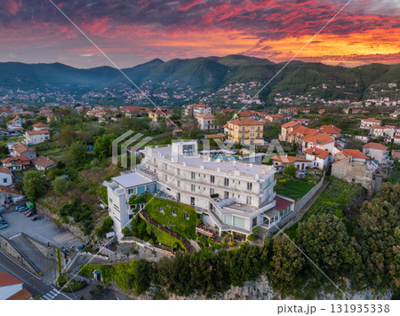 Aerial view of a luxury hotel in Amalfi, Italy, with terraces, a rooftop pool, red tiled houses, lush greenery, and a vibrant sunset sky. 131935338