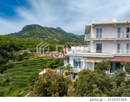 Luxury hotel with white facade on a hillside in Amalfi, Italy, surrounded by greenery, terraced fields, rolling hills, and a partly cloudy sky. Luxury hotel with white facade on a hillside in Amalfi, Italy, surrounded by greenery, terraced fields, rolling hills, and a partly cloudy sky. 131935369