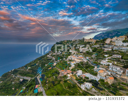 Aerial view of Ravello, Italy, with pastel buildings, terraced gardens, and the Mediterranean Sea under a vibrant pink and orange sky. 131935372