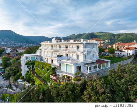 A multi story white building with balconies and rooftop area on a hillside, surrounded by greenery, with red roofed town and hills in the distance. 131935382