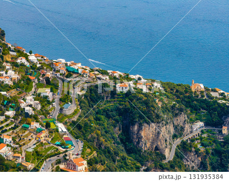 Aerial view of Amalfi, Italy, showing colorful buildings on green hills, a cliff with a tunnel, a church bell tower, and the Tyrrhenian Sea with a boat. 131935384