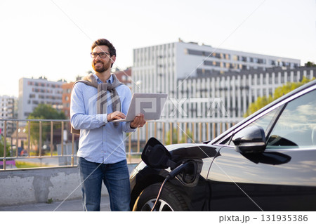 A man stands next to his electric vehicle while using a laptop, possibly monitoring the charging process. 131935386