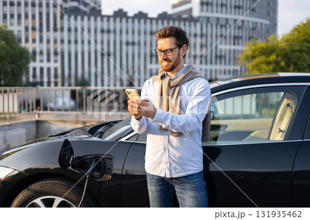 A man checks his phone while his electric car charges at a charging station in an urban setting. A man checks his phone while his electric car charges at a charging station in an urban setting. 131935462