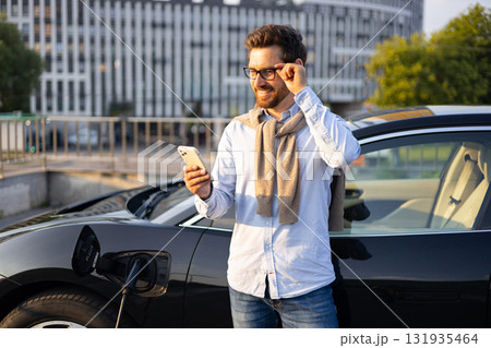 A man checks his phone while his electric vehicle charges at a charging station in an urban setting. A man checks his phone while his electric vehicle charges at a charging station in an urban setting. 131935464