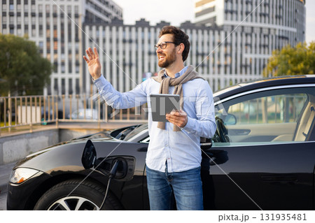 A man waves while standing next to his electric vehicle, which is charging at a station in an urban setting. A man waves while standing next to his electric vehicle, which is charging at a station in an urban setting. 131935481
