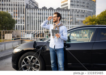 A man stands next to his electric car while it charges, holding a tablet and looking off into the distance. A man stands next to his electric car while it charges, holding a tablet and looking off into the distance. 131935485