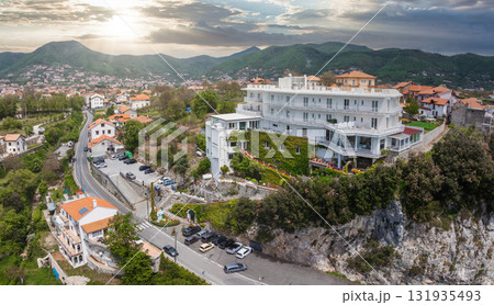 Aerial view of a white ivy covered hotel on a rocky cliff, surrounded by greenery, a winding road, terracotta roofed houses, and rolling hills at sunset. Aerial view of a white ivy covered hotel on a rocky cliff, surrounded by greenery, a winding road, terracotta roofed houses, and rolling hills at sunset. 131935493