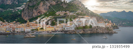 Aerial view of Amalfi, Italy, showing colorful buildings on cliffs, a church tower, lush hills, a hillside hotel, and boats on the Tyrrhenian Sea. 131935499