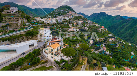 Aerial view of Ravello, Italy, featuring white and pastel buildings, terraced gardens, a modern structure, and lush green mountains under a cloudy sky. Aerial view of Ravello, Italy, featuring white and pastel buildings, terraced gardens, a modern structure, and lush green mountains under a cloudy sky. 131935517