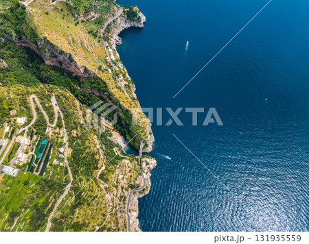 Aerial image of the Amalfi Coast, Italy, featuring cliffs, winding roads, terraced fields, green hills, and a boat on the Tyrrhenian Sea. Aerial image of the Amalfi Coast, Italy, featuring cliffs, winding roads, terraced fields, green hills, and a boat on the Tyrrhenian Sea. 131935559