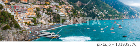 Positano on Italy's Amalfi Coast features vibrant buildings on steep cliffs, a busy beach with umbrellas, a ferry, and boats in turquoise waters. 131935576