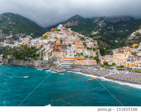 Positano on Italy's Amalfi Coast features vibrant hillside buildings, a sandy beach with umbrellas, turquoise waters, and lush mountains with clouds. Positano on Italy's Amalfi Coast features vibrant hillside buildings, a sandy beach with umbrellas, turquoise waters, and lush mountains with clouds. 131935586