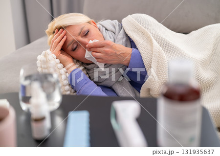Blonde woman lying on sofa at home, feeling weak with runny nose and fever. Medicines and thermometer placed on table in front. Concept of illness, flu, and home treatment. Blonde woman lying on sofa at home, feeling weak with runny nose and fever. Medicines and thermometer placed on table in front. Concept of illness, flu, and home treatment. 131935637