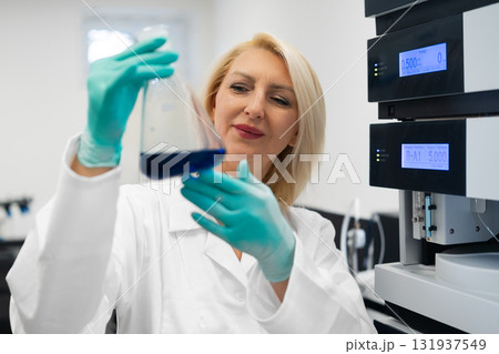 Blonde female scientist in rubber gloves inspecting a liquid microbial culture in a flask while standing in a microbiology laboratory with HPLC and lab equipment in the background 131937549
