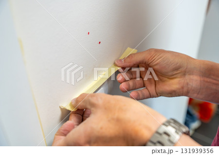 Man covering a light switch with masking tape before painting the wall white. Home renovation, surface protection and careful preparation during interior work. Man covering a light switch with masking tape before painting the wall white. Home renovation, surface protection and careful preparation during interior work. 131939356