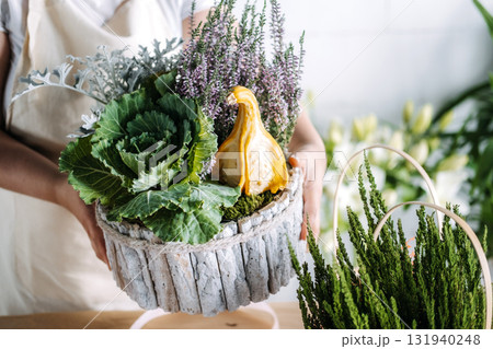Person holding a finished botanical centerpiece featuring cabbage, dusty miller, heather, and a yellow gourd. Thanksgiving table decor, seasonal centerpiece, botanical tablescape, holiday arrangement Person holding a finished botanical centerpiece featuring cabbage, dusty miller, heather, and a yellow gourd. Thanksgiving table decor, seasonal centerpiece, botanical tablescape, holiday arrangement 131940248