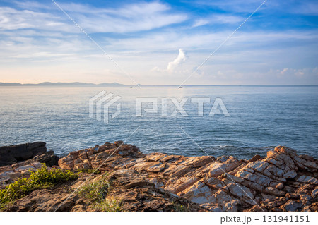 Striking coastal rock formations at Khao Laem Ya, Rayong, Thailand, contrasting with the calm blue ocean and wispy clouds under a bright summer sky 131941151