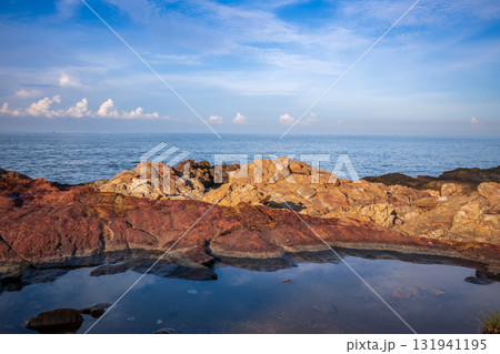 Close-up of rugged red and golden rocks at Khao Laem Ya, Rayong, Thailand, with a tidal pool reflection and calm blue sea under a bright sky Close-up of rugged red and golden rocks at Khao Laem Ya, Rayong, Thailand, with a tidal pool reflection and calm blue sea under a bright sky 131941195