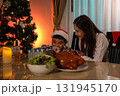 Curious boy opening Christmas gift box with his loving mother beside decorated tree at home. Warm holiday atmosphere with festive dinner, candlelight, and joyful family celebration full of excitement 131945170