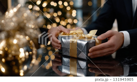 Man in suit giving gift box with gold ribbon, celebrating holiday presenting luxury present on reflective surface with festive bokeh lights in background, formal occasion 131945905